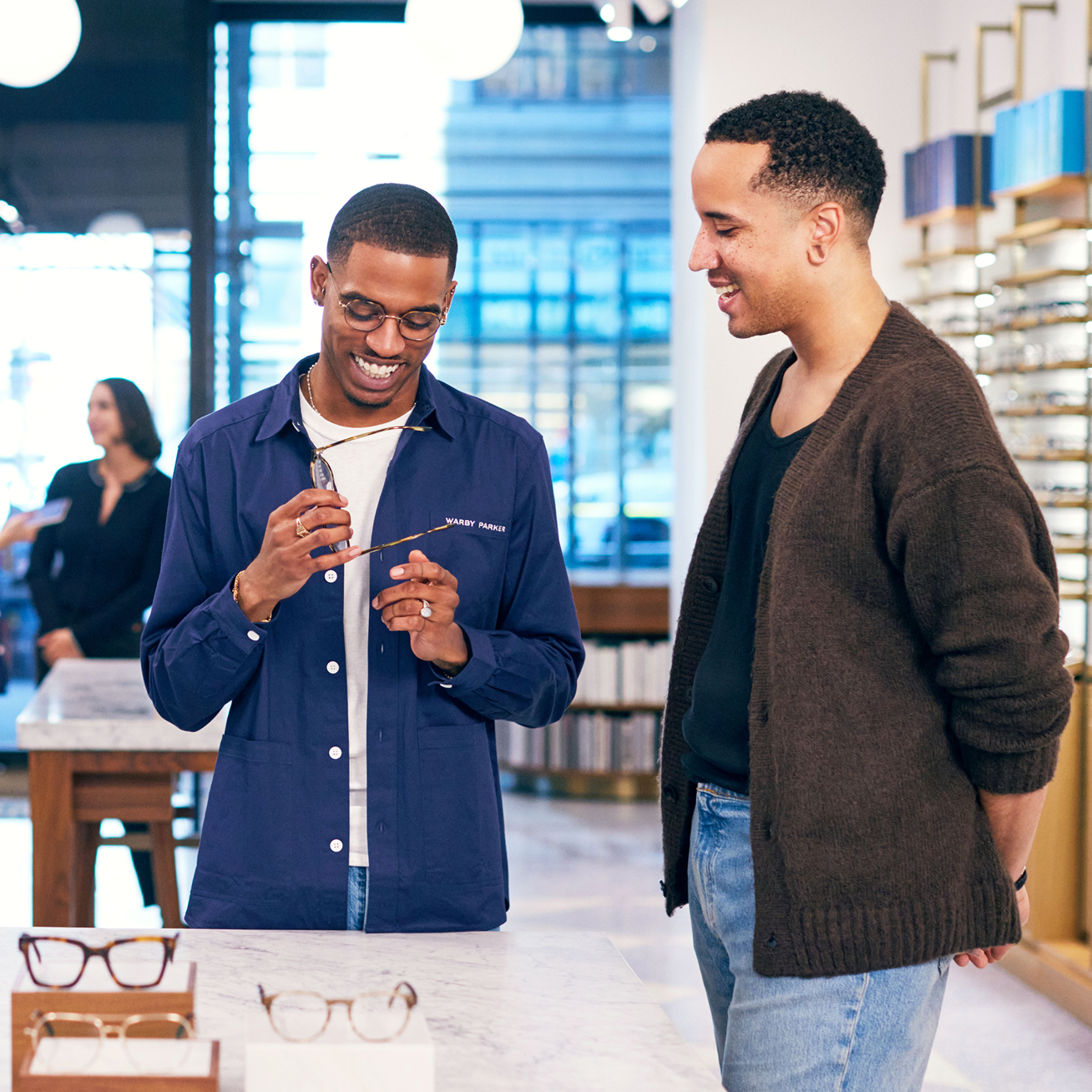 Two men look at glasses in a store. One man holds up a pair of glasses to his face, and the other looks on.