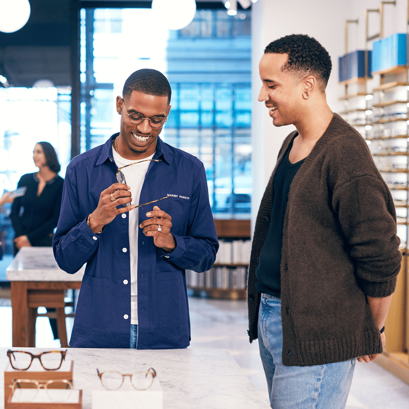 Two men look at glasses in a store. One man holds up a pair of glasses to his face, and the other looks on.