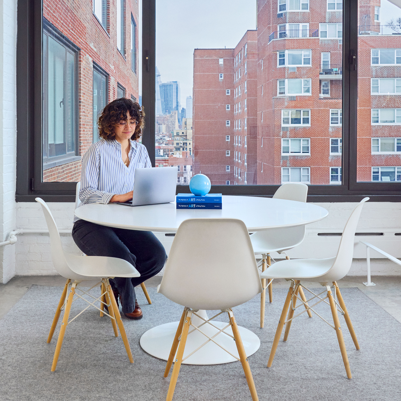 A woman sits at a round white table with a laptop, with an urban backdrop.