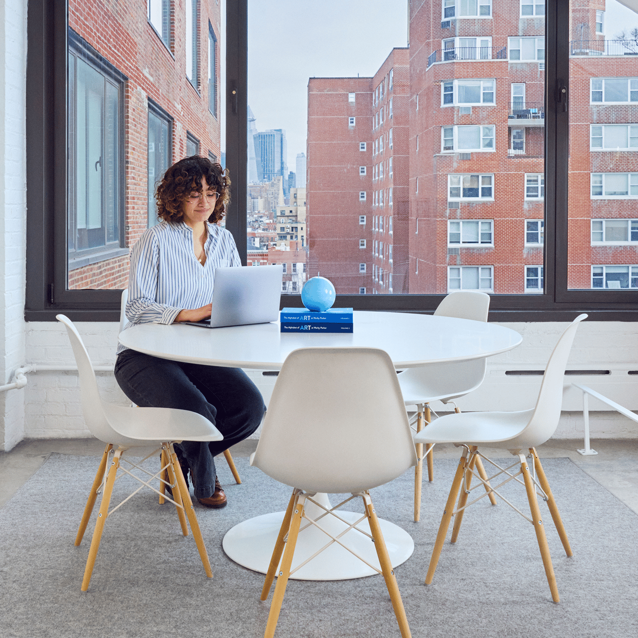 A woman sits at a round white table with a laptop, with an urban backdrop.