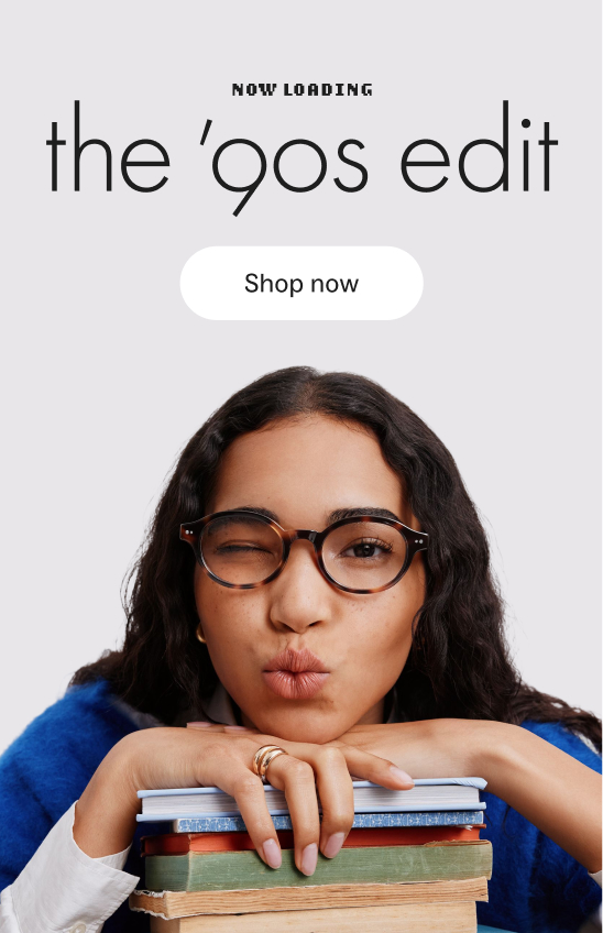 A woman wearing glasses and a blue sweater winks while leaning on a stack of books.