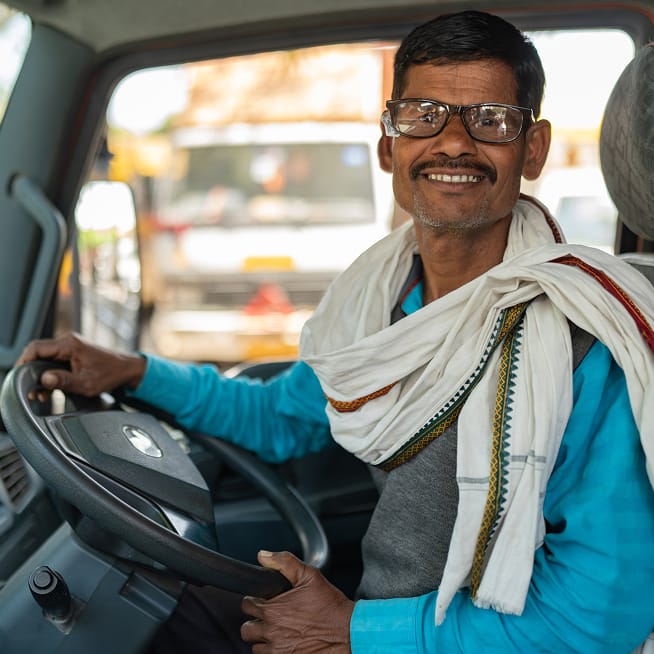 Smiling driver wearing glasses and a scarf, sitting in a vehicle's driver seat.
