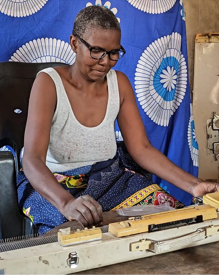 A woman wearing glasses is focused on using a knitting machine. She sits against a blue patterned fabric background.