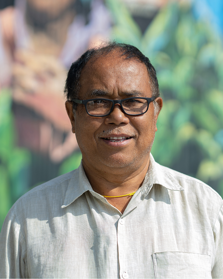 A smiling man wearing glasses and a light-colored shirt stands outdoors with a colorful mural backdrop.