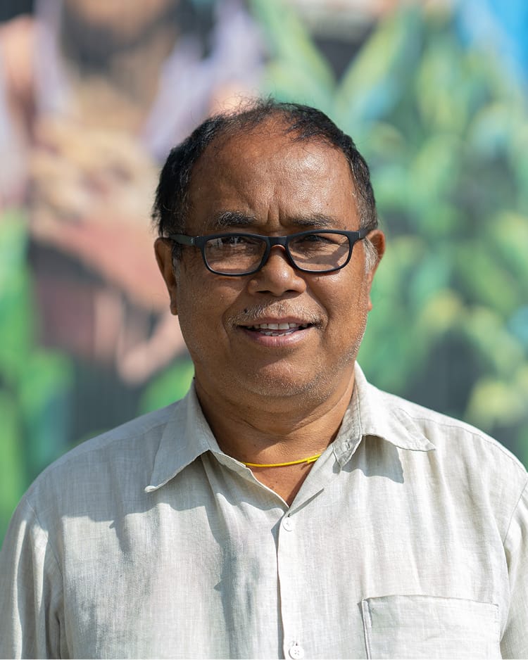 A smiling man wearing glasses and a light-colored shirt stands outdoors with a colorful mural backdrop.
