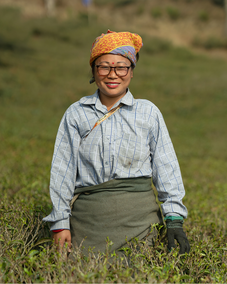 A smiling woman wearing glasses, a patterned headscarf, and a checked shirt stands in a lush green field.