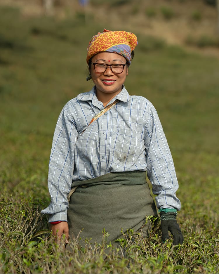 A smiling woman wearing glasses, a patterned headscarf, and a checked shirt stands in a lush green field.