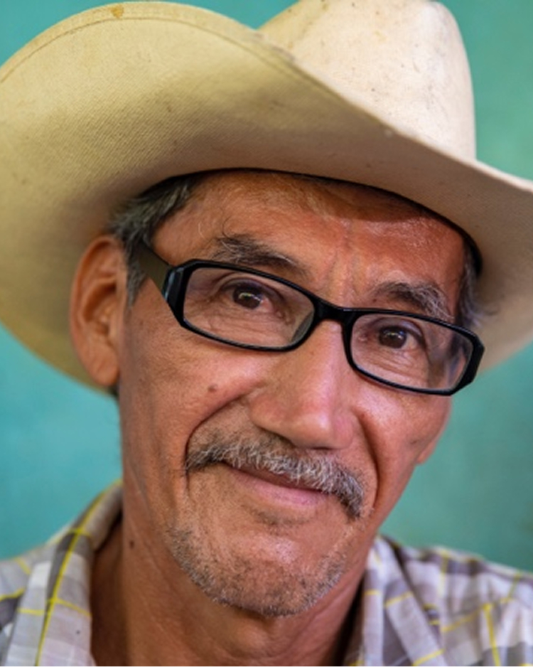 A man wearing glasses and a cowboy hat smiles, with a light blue background.