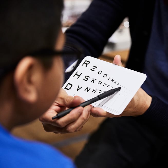 Eye exam with chart; smiling girl with glasses and peace sign in front of a bookshelf.