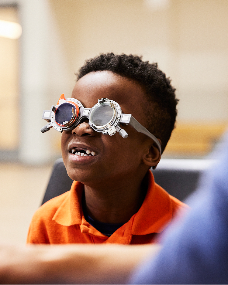 A child wearing an orange shirt tries on a pair of trial lenses, smiling in an indoor setting.