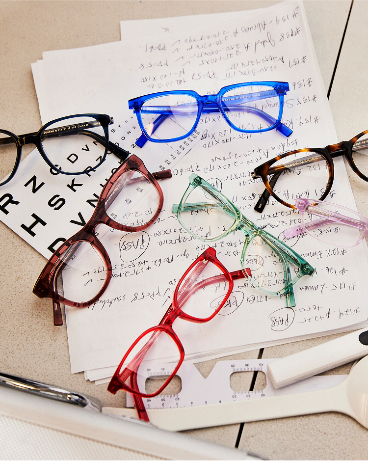 Colorful eyeglasses are scattered on handwritten notes and an eye chart.