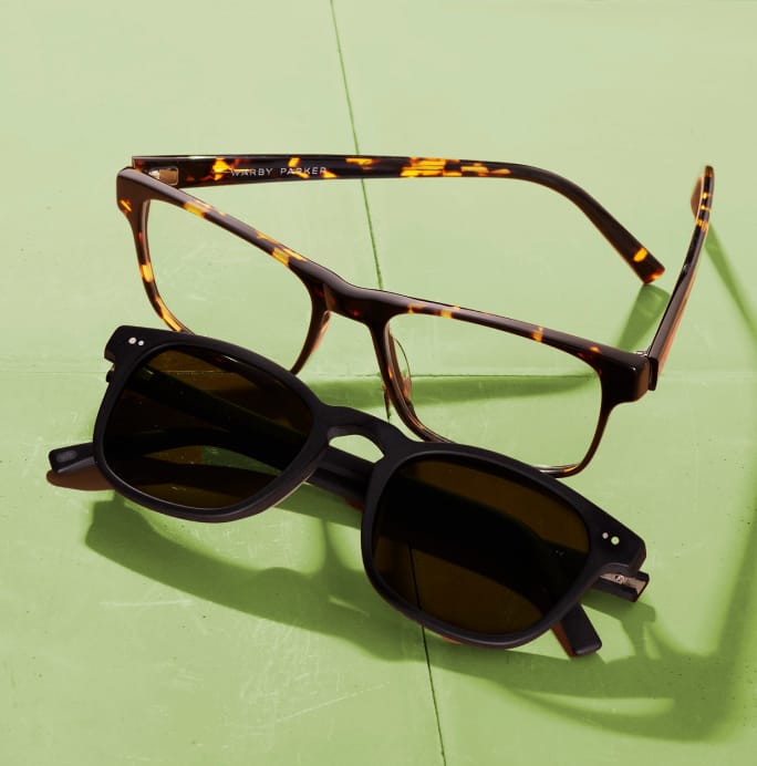 A pair of tortoiseshell glasses is stacked on top of a pair of black sunglasses on a light green surface.