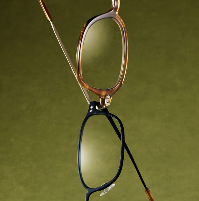 Two pairs of glasses, one tortoise-shell and one black, are hanging and overlapping on a green background.