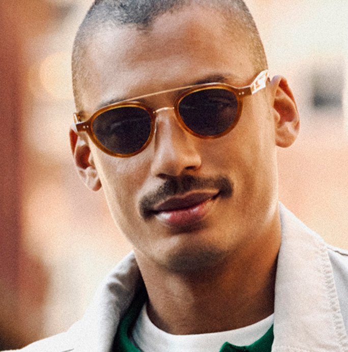 Close-up of a man with a mustache and short haircut wearing sunglasses and a light-colored jacket.