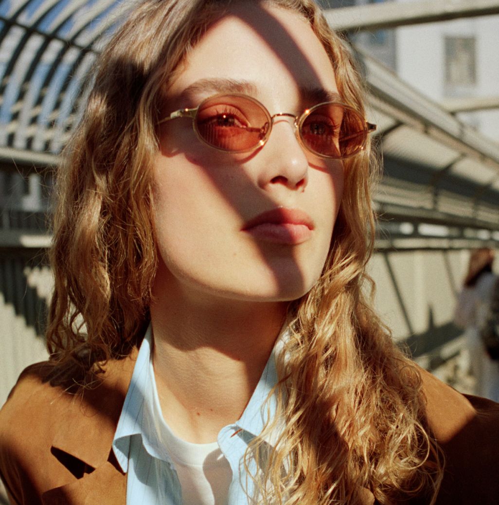 A young woman with wavy blonde hair and a serious expression wears rose-tinted oval sunglasses and a tan jacket.