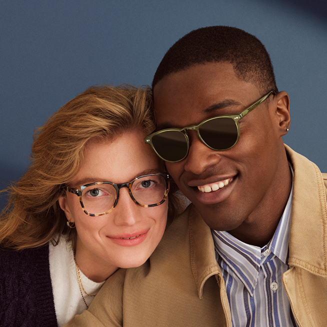 Two people wearing stylish eyeglasses, one with round gold frames and the other with tortoiseshell frames, posing together.