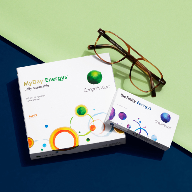 Two CooperVision contact lens boxes and brown glasses on green and blue background.