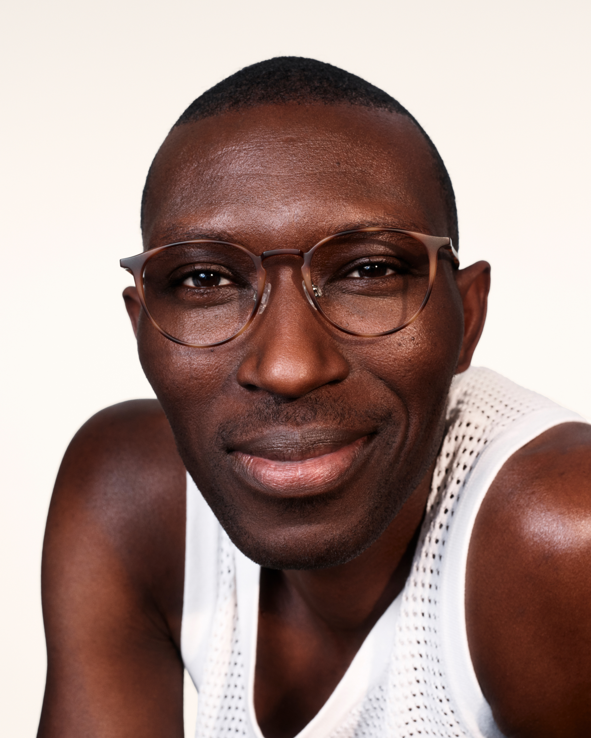 A man wearing glasses and a white tank top smiles into the camera.