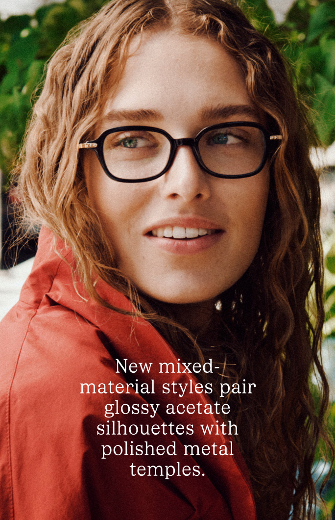 A woman with curly auburn hair and glasses smiles as she drinks from a white mug.