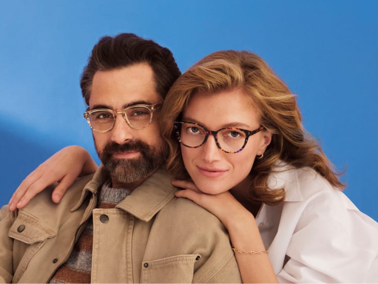 Man and woman wearing glasses posing closely against a blue background.