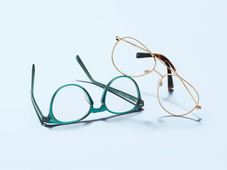 Pair of teal round glasses and pair of gold aviator glasses on a light blue surface.