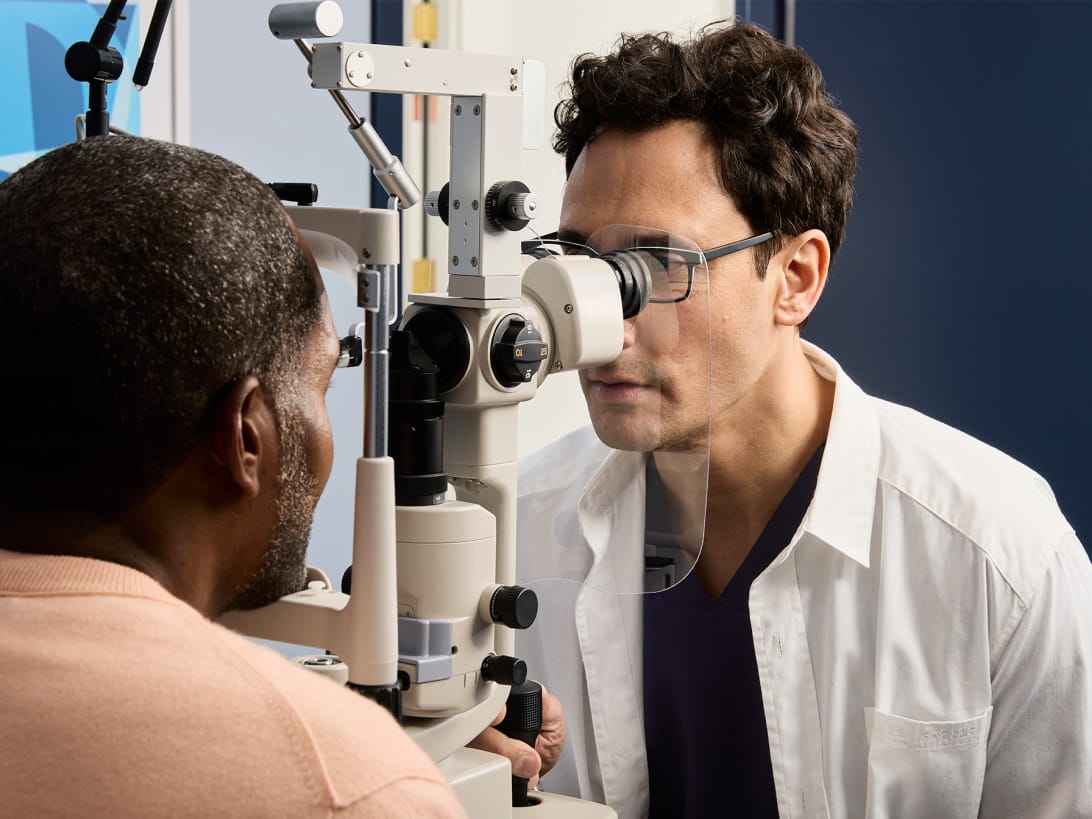 Male eye doctor wearing glasses and a white coat examining a male patient’s eyes using a slit lamp in a medical office.