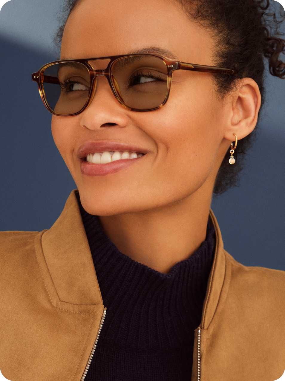 Person with curly hair wearing sunglasses and a brown jacket.