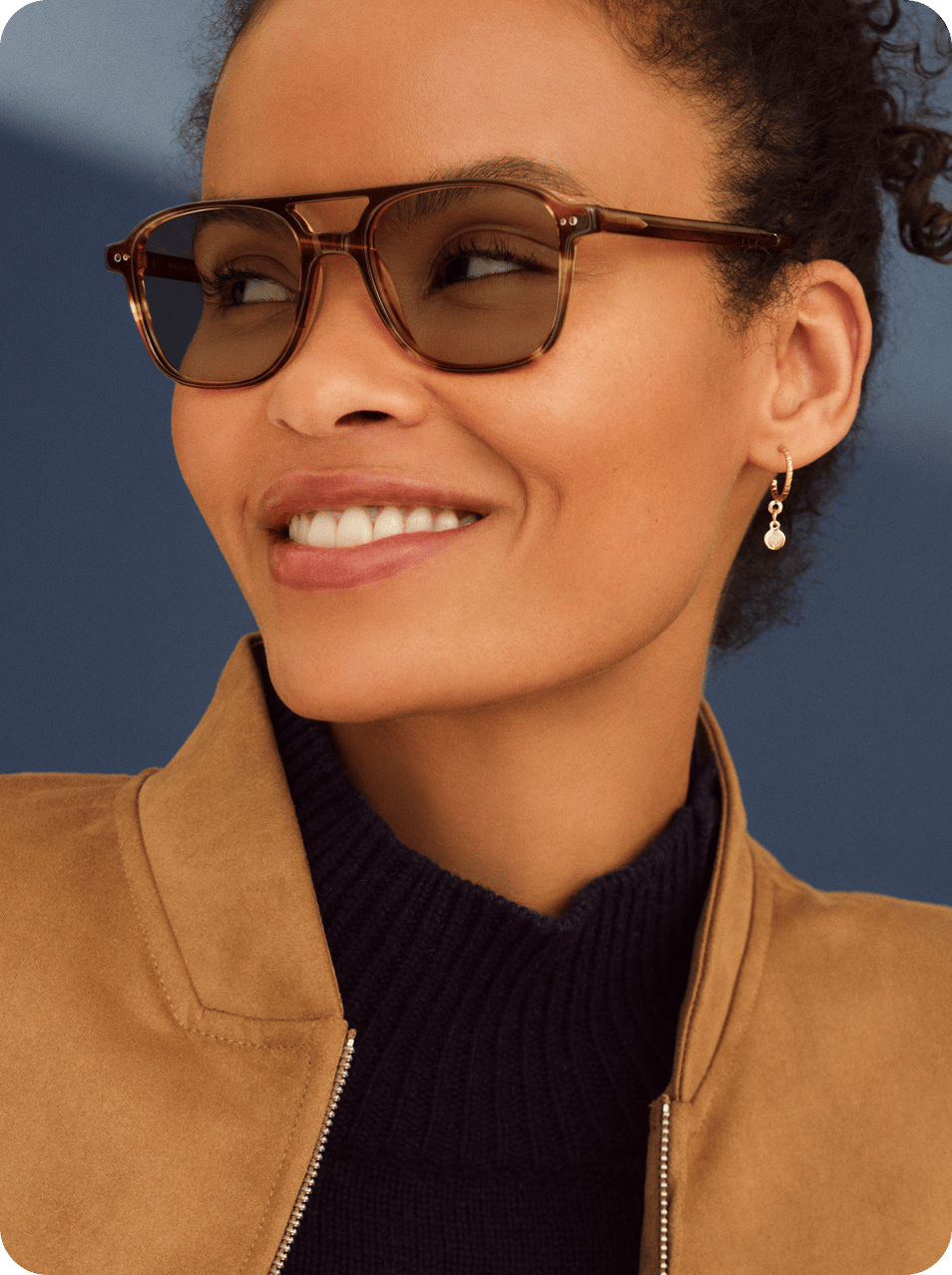 Person with curly hair wearing sunglasses and a brown jacket.